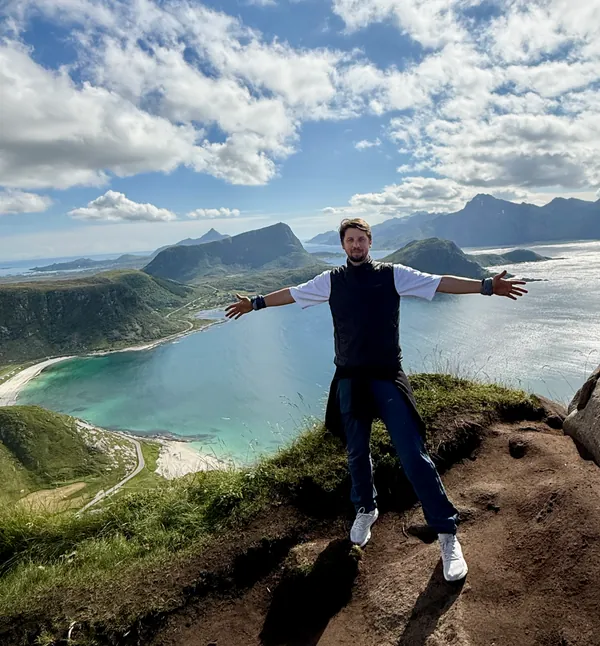 Håkon Ege standing with arms outstretched above a mountainous coastline.